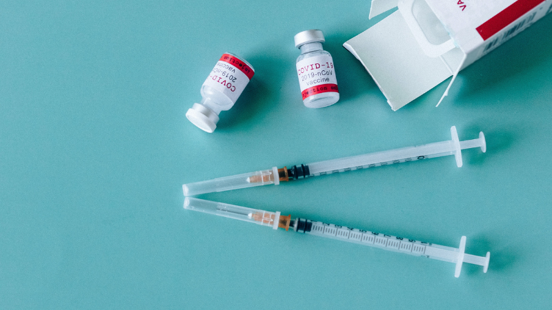 Light blue background, two vaccine needles, two COVID-19 vaccine doses in small glass bottles, and an open box of vaccines. Photo by Nataliya Vaitkevich.